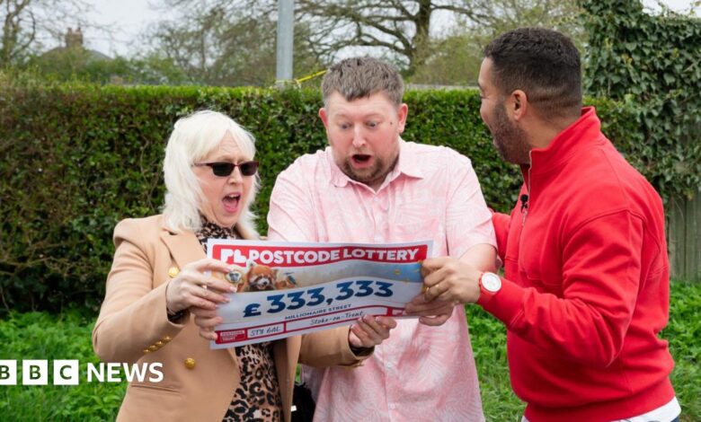 A group of three people standing outdoors. One is wearing a red jumper while the other two are wearing a brown coat and pink shirt and holding a piece of paper which has been styled to look like a cheque.