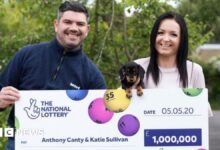Anthony Canty is wearing a navy quarter zip jumper. He has dark hair and is holding a large cheque for £1m outdoors. Also holding the cheque, and a small black dog, is his partner, Katie Sullivan. She has long dark hair. Both are smiling.