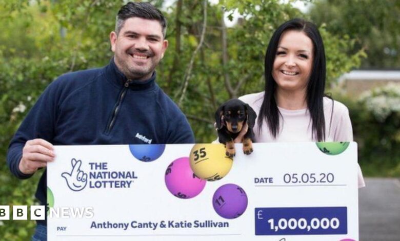 Anthony Canty is wearing a navy quarter zip jumper. He has dark hair and is holding a large cheque for £1m outdoors. Also holding the cheque, and a small black dog, is his partner, Katie Sullivan. She has long dark hair. Both are smiling.