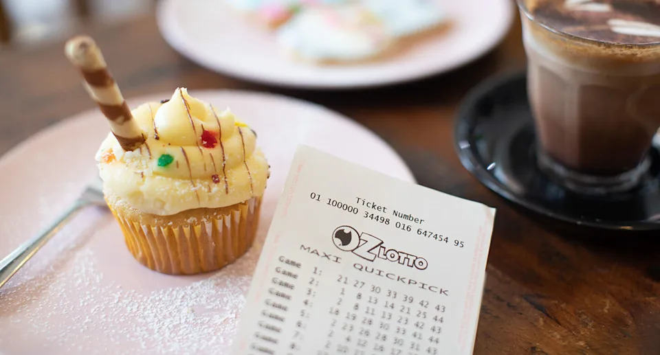 An Oz Lotto ticket sits on a plate next to a cupcake and a coffee.