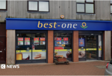 A shop with the words Best One on a blue background at the top of the ground floor outside. Part of a pavement and a silver car are visible in front of the store.