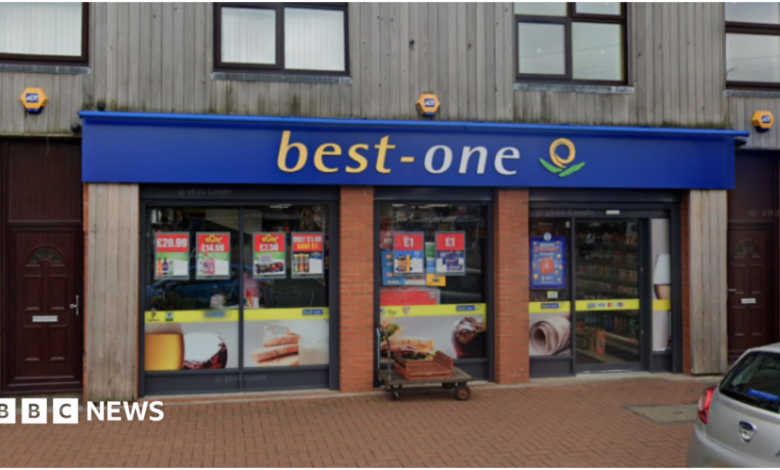 A shop with the words Best One on a blue background at the top of the ground floor outside. Part of a pavement and a silver car are visible in front of the store.