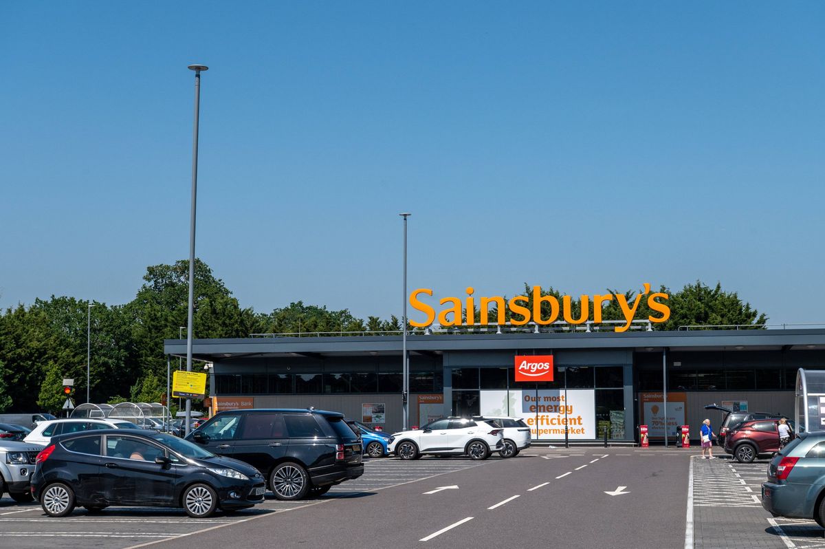 An image depicts a parking lot in front of a supermarket with multiple vehicles parked, including cars and SUVs, with a clear sky and a row of trees in the background.