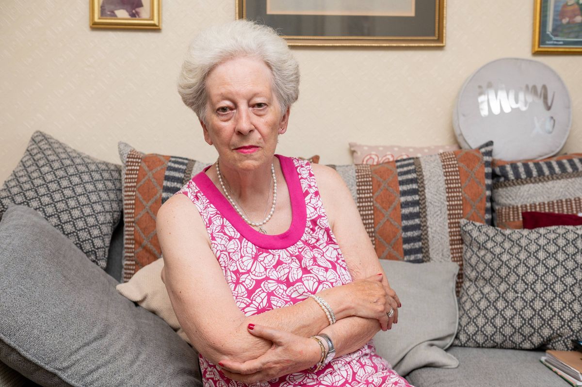 An elderly individual, dressed in a pink sleeveless dress with a white trim, is seated on a cushioned sofa, her arms crossed and her gaze directed downward, in a living room adorned with various decorative pillows and framed artwork on the walls.