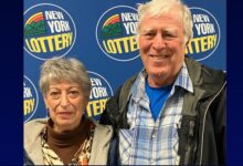 An older man smiles next to a woman, both standing in front of a blue New York Lottery logo backdrop.
