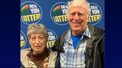 An older man smiles next to a woman, both standing in front of a blue New York Lottery logo backdrop.