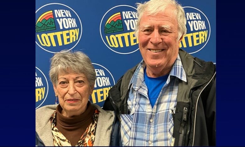 An older man smiles next to a woman, both standing in front of a blue New York Lottery logo backdrop.