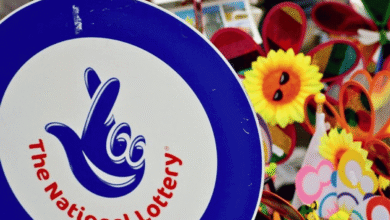 A sign featuring "The National Lottery" logo with a blue background and a hand symbol with crossed fingers. In the background, there are colorful pinwheels and sunflowers with smiling faces, likely part of a festive street stall or market. UK's National Lottery to temporarily shut down for major tech updates. UK's National Lottery returns to '90s 'It Could Be You' tagline. Court of Appeal blocks Richard Desmond companies National Lottery license challenge