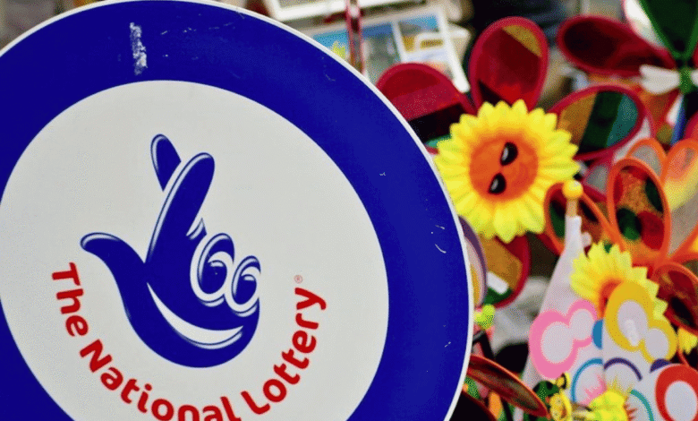 A sign featuring "The National Lottery" logo with a blue background and a hand symbol with crossed fingers. In the background, there are colorful pinwheels and sunflowers with smiling faces, likely part of a festive street stall or market. UK's National Lottery to temporarily shut down for major tech updates. UK's National Lottery returns to '90s 'It Could Be You' tagline. Court of Appeal blocks Richard Desmond companies National Lottery license challenge