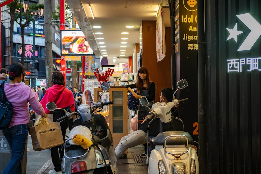 People walk past scooters parked near the entrance of a shop.