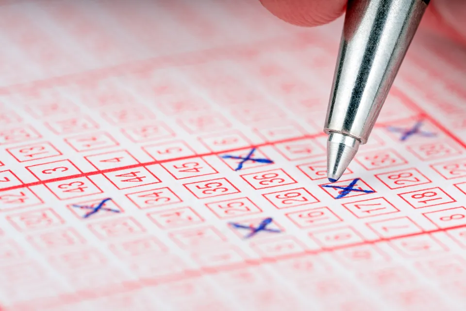 Close-up of a hand with a silver pen marking numbers on a red-lined lottery ticket.