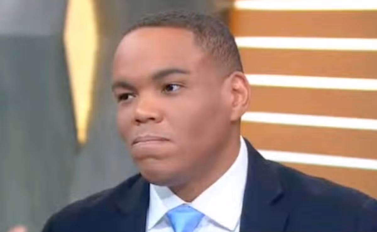 An individual in formal attire, comprising a dark suit jacket and a light blue tie, stands attentively in front of a backdrop featuring a combination of vertical stripes and a geometric pattern, possibly during an interview or public speaking engagement.